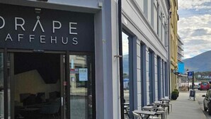 Rakennuksessa on myymälätila, jonka yläpuolella on kyltti, jossa lukee ”DRAPE AFFEHUUS”, ja sen edessä jalkakäytävällä on ulkoilmakalusteita.