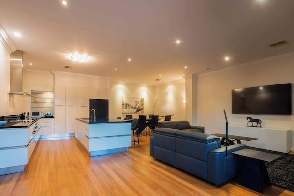A modern living room with a sofa, a dining area, a kitchen, and a TV mounted on the wall.