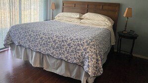 A bedroom with a large bed, two bedside lamps, a framed floral artwork on the wall, and a window with sheer curtains.