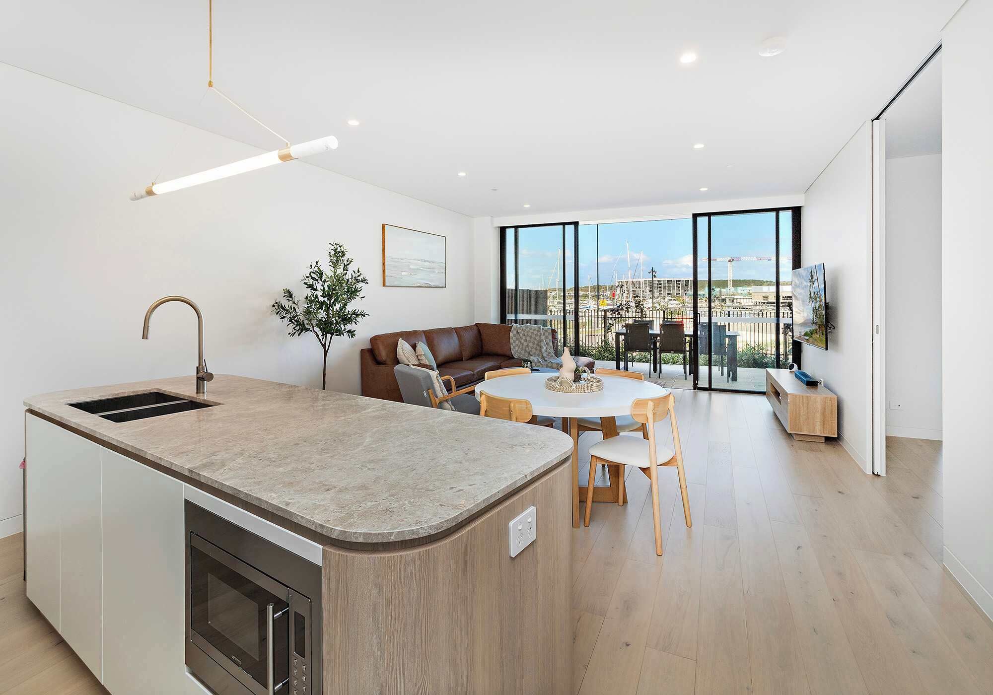A modern kitchen with a built-in oven, a dining area with a round table and chairs, a sofa, and a large window offering a city view.