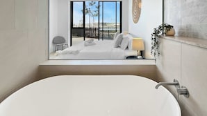 Un baño moderno con una bañera grande, una mesita de noche pequeña con lámpara, y vistas a un balcón con cama y sillón.