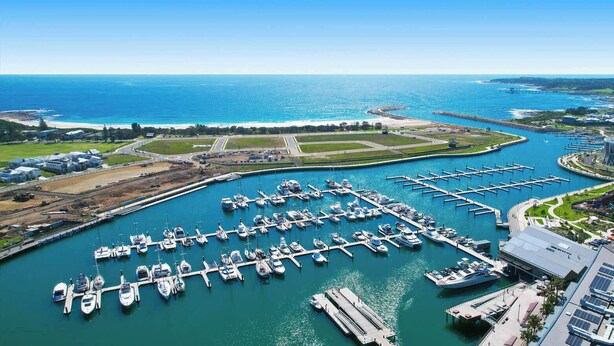 Un puerto deportivo con numerosas embarcaciones amarradas, un edificio grande con paneles solares y una vista despejada del océano y la costa.