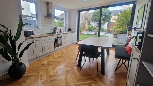A modern kitchen with white cabinets, a wooden herringbone floor, a dining table with chairs, and a large window offering a view of the outdoors.