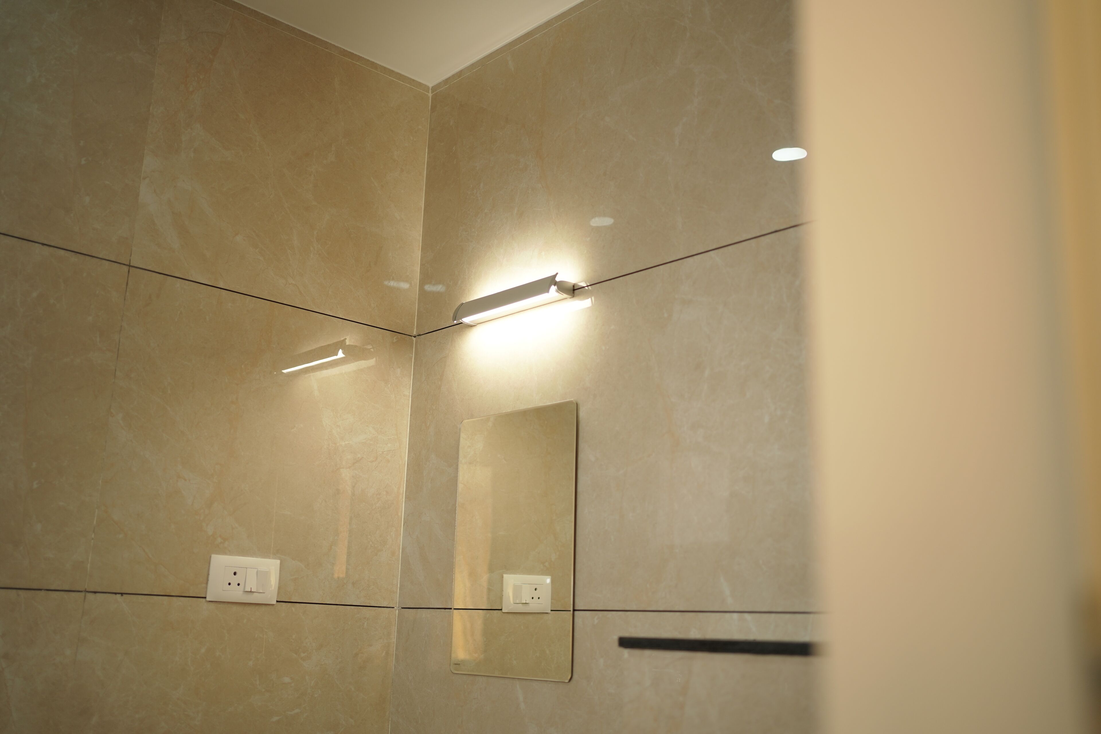 Un baño con paredes de mármol color crema, un espejo y una lámpara sobre el espejo.