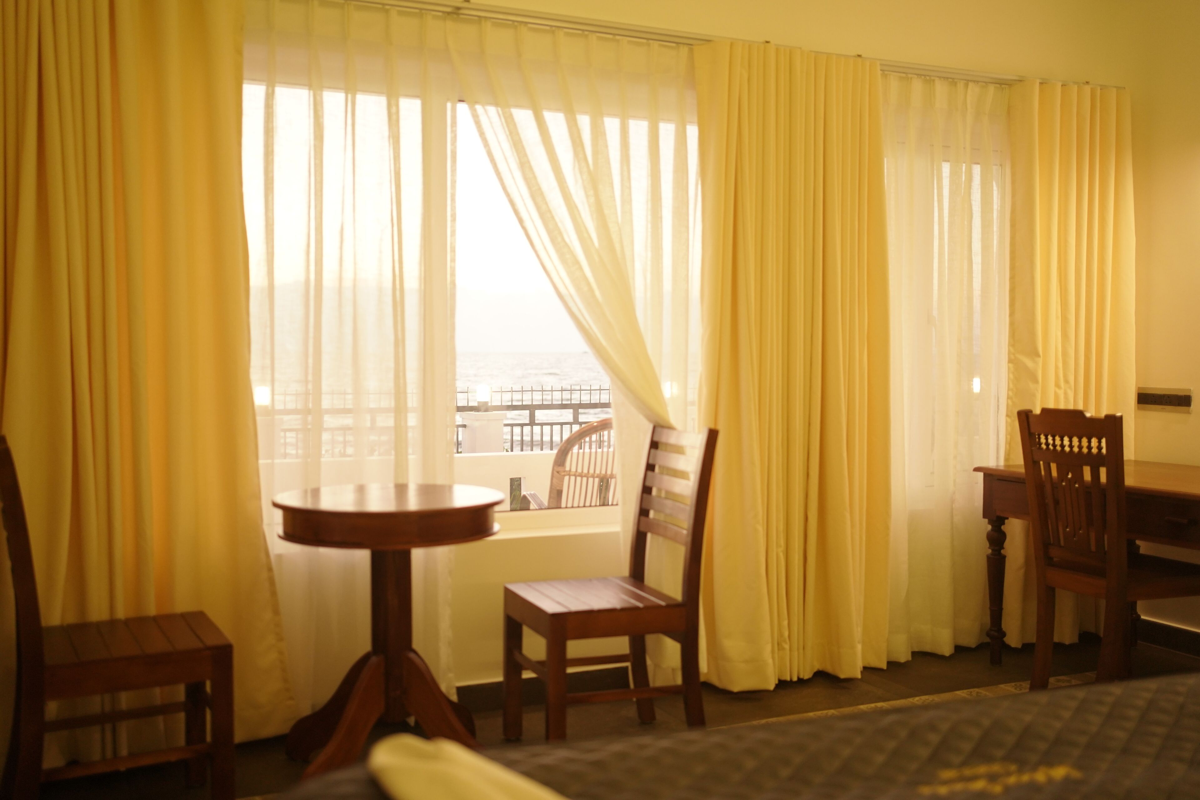A room with a wooden chair, a small round table, and a view through a window with yellow curtains.