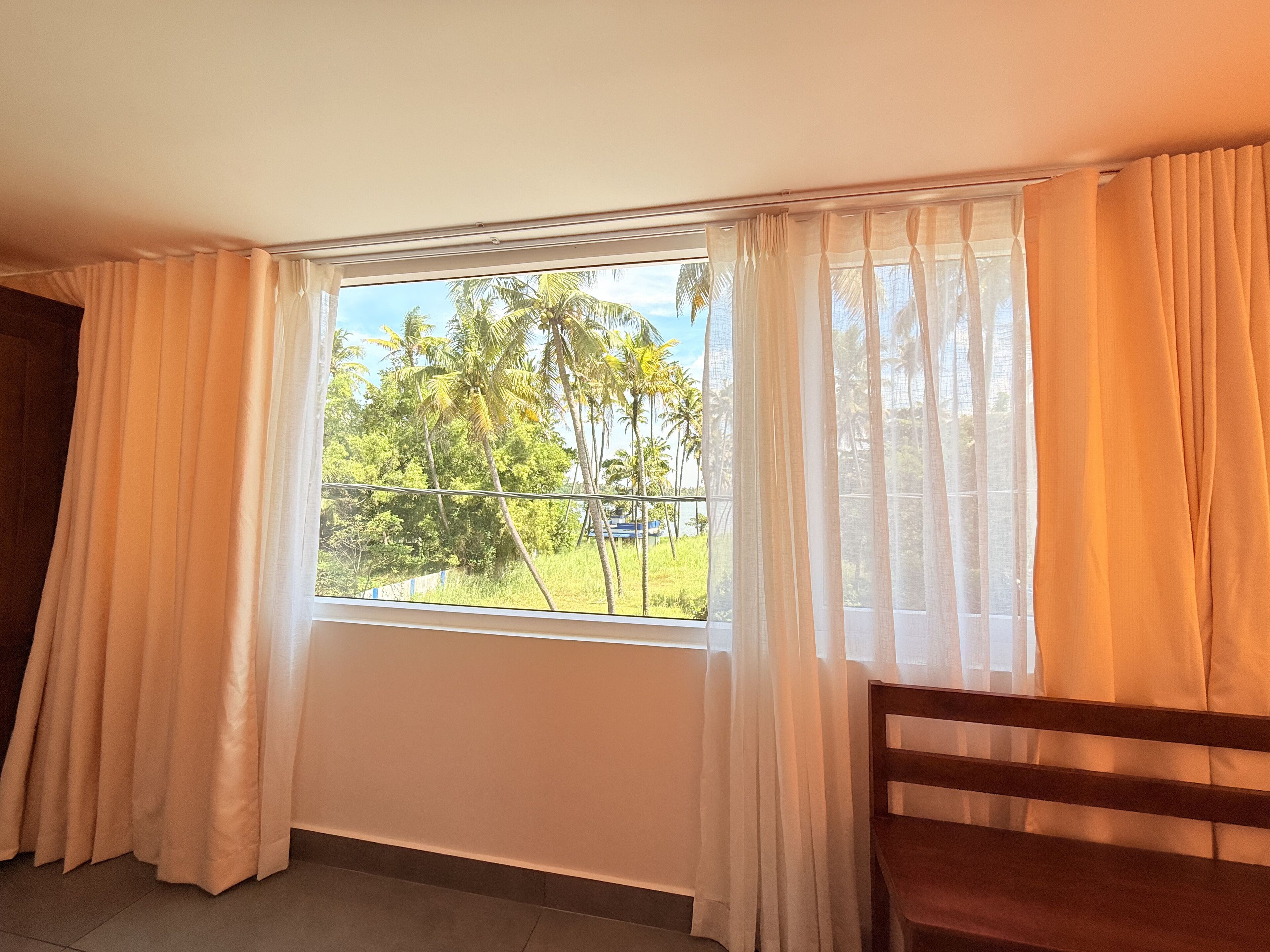 A room with a large window, a wooden chair, and a view of palm trees and greenery outside.