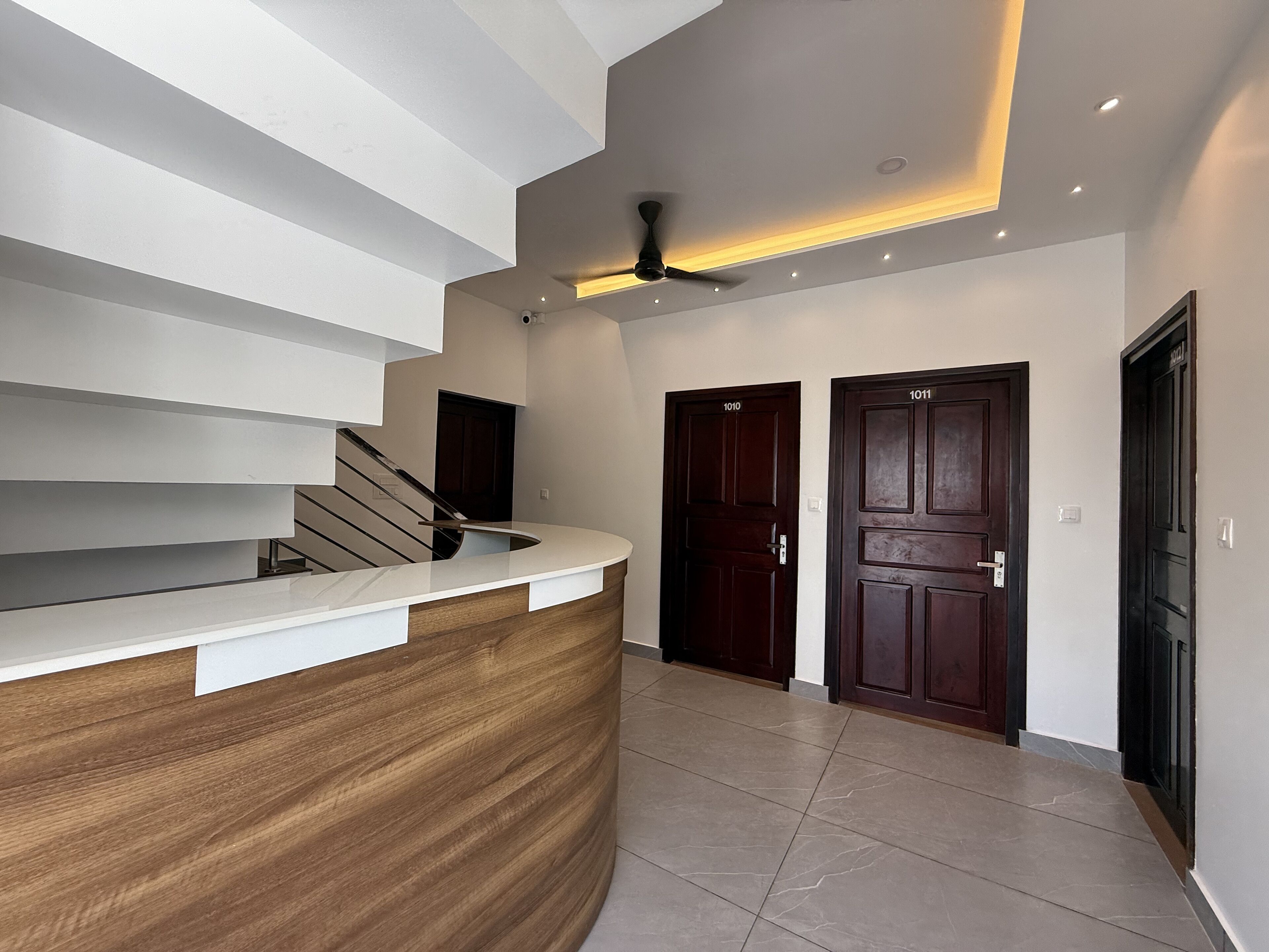 Un espace d’accueil moderne avec un comptoir en bois aux lignes courbes, un escalier et deux portes en bois foncé portant les numéros 1010 et 1011.