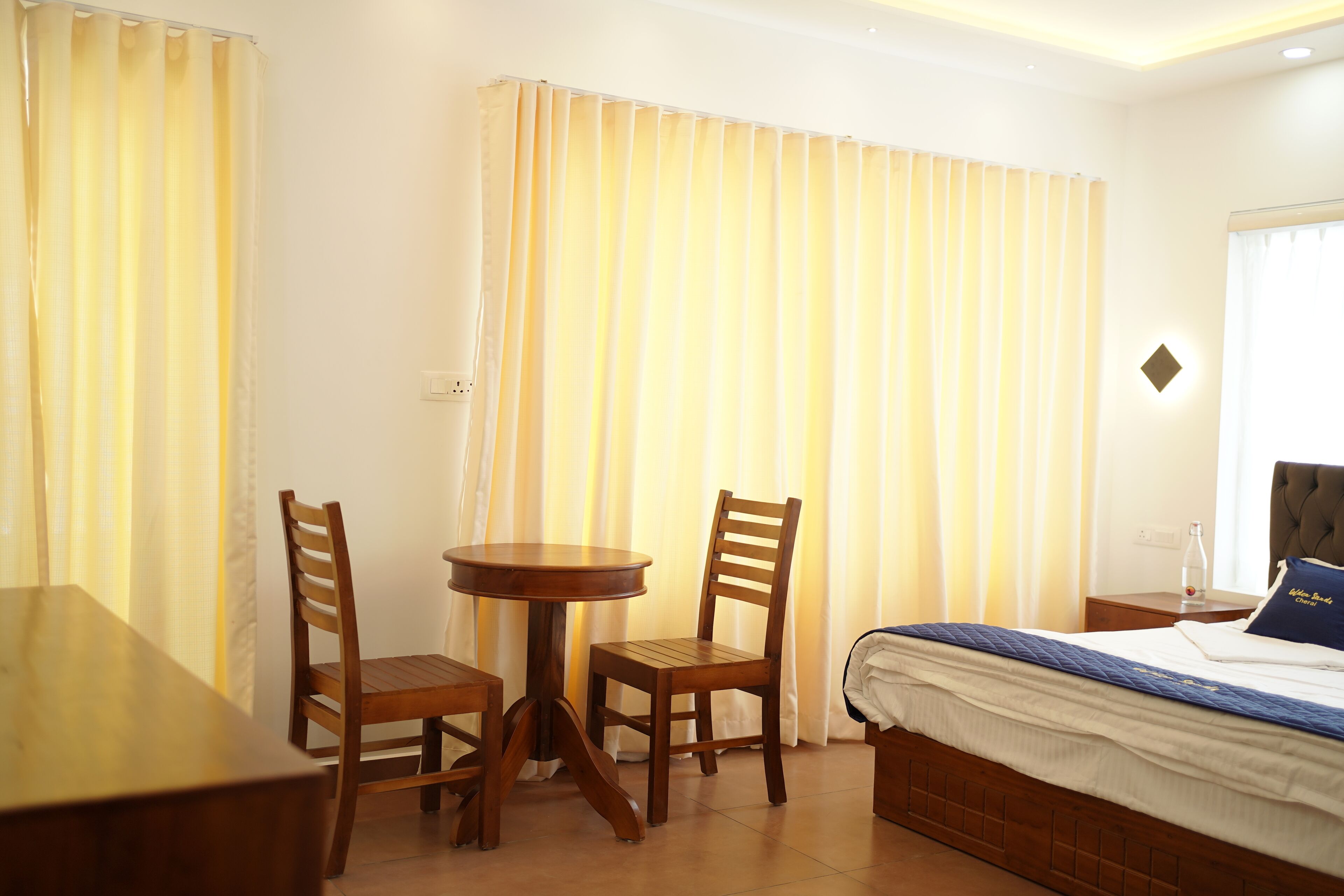 A hotel room with a bed, a wooden table with two chairs, and a window with curtains.