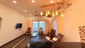 A spacious living room with a large black table, two wooden chairs, a ceiling fan, and a modern chandelier. - Golden Sands Cherai (Kochi)