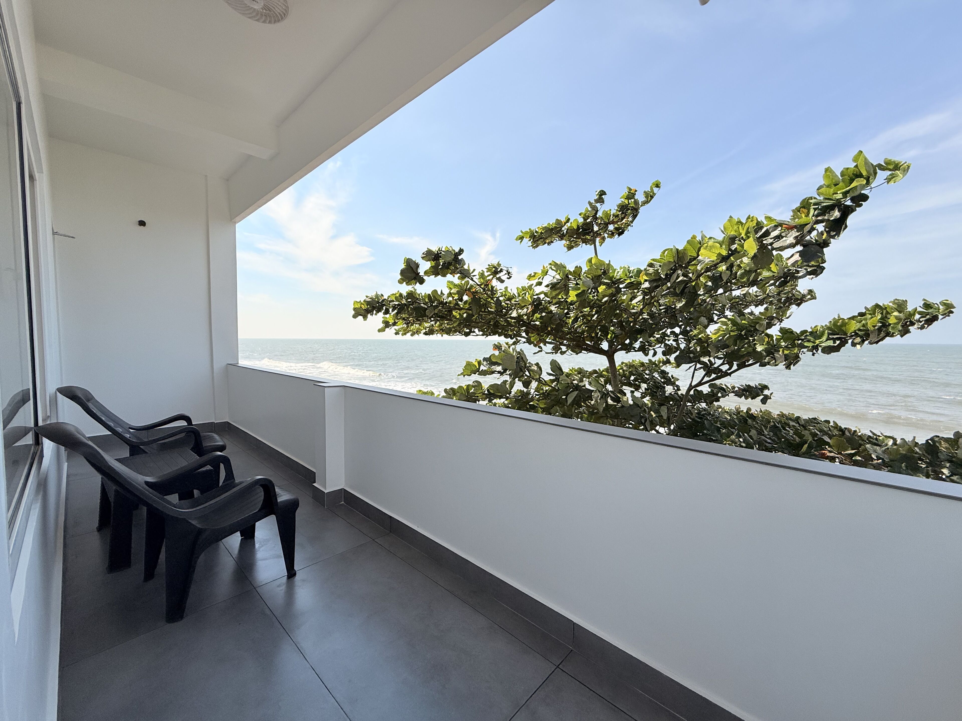 A balcony with a view of the sea, a tree, and two black lounge chairs.