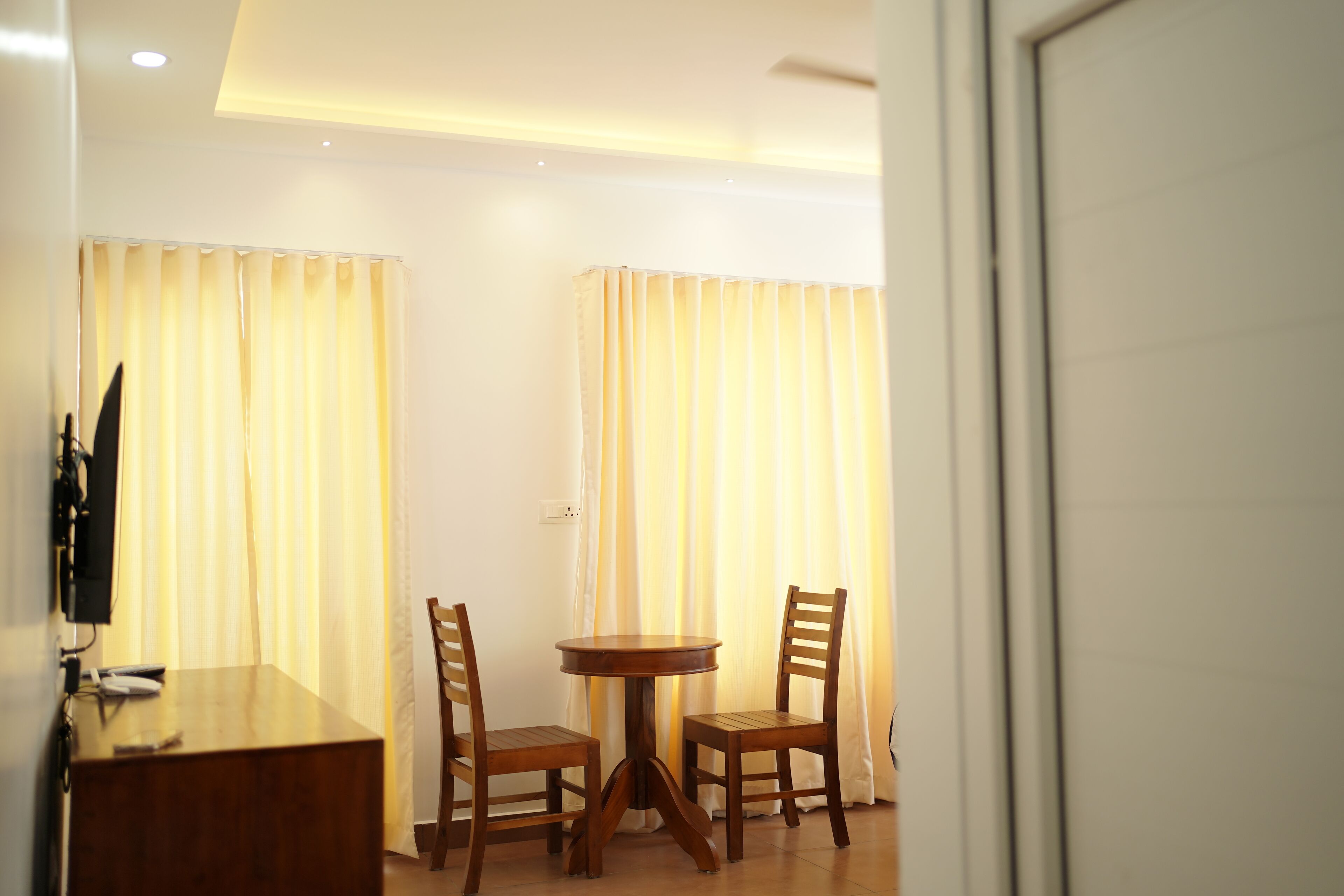 A room with a wooden table and two chairs, a TV on the wall, and curtains on the windows.