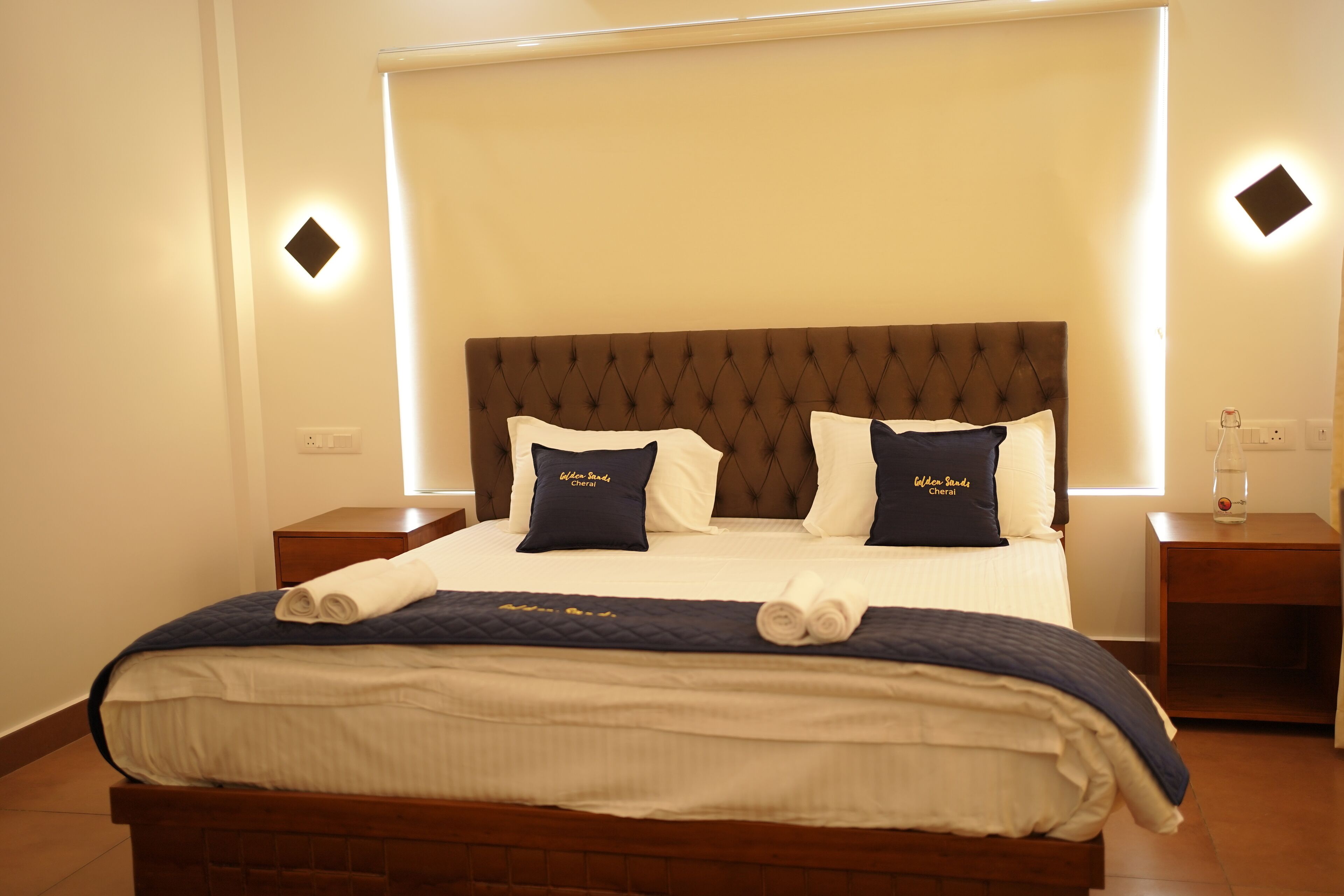 A neatly made bed with a dark headboard, two pillows with text, and a rolled towel on the bed. There are two bedside tables with a bottle on one.