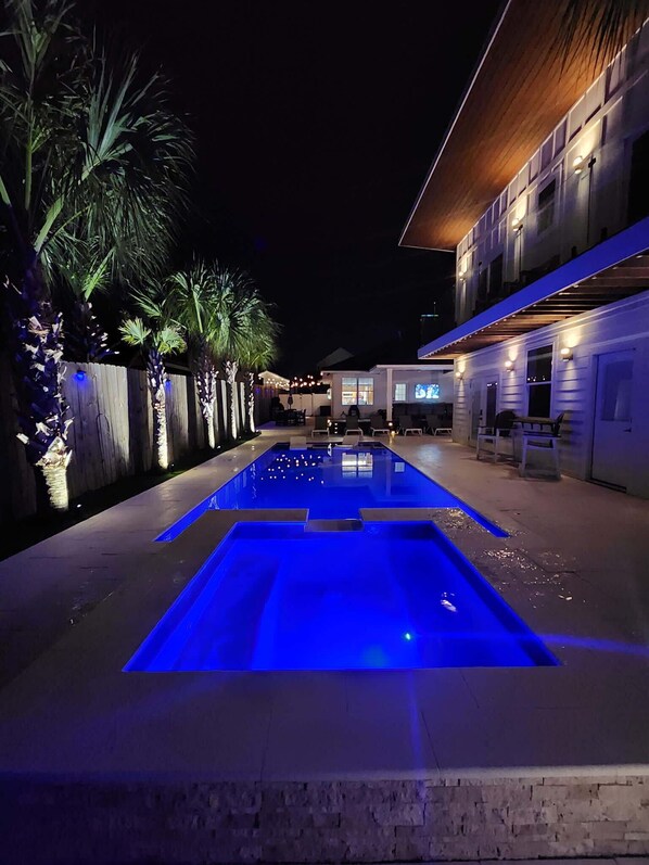 A swimming pool with blue lighting, surrounded by a wooden fence and palm trees.