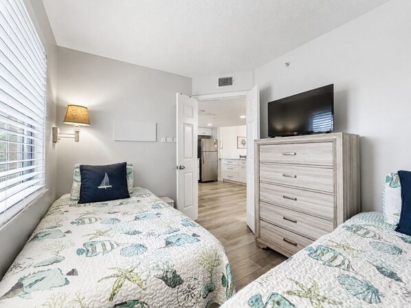 A bedroom with two beds, a dresser, a television, and a hallway leading to a kitchen.