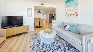 A modern living room with a flat-screen TV, a white sofa, a coffee table, and a patterned rug. - Corner Beachfront 2BR | Balcony & Pool (St. Pete Beach)