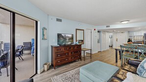 A spacious living room with a large TV, wooden furniture, and a view through a sliding glass door. - Coastal Beachfront Condo | Pool & Jacuzzi (St. Pete Beach)