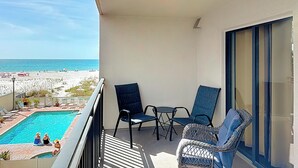 A balcony with a view of the beach, two wicker chairs, a small table, and a pool. - Coastal Beachfront Condo | Pool & Jacuzzi (St. Pete Beach)