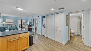 A modern kitchen with wooden cabinets, a dining area with blue chairs, and a hallway leading to a bathroom. - Coastal Beachfront Condo | Pool & Jacuzzi (St. Pete Beach)
