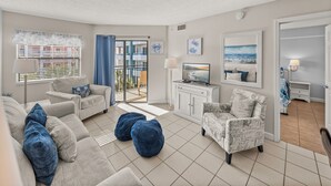 Ein Wohnzimmer mit einer Couch, Sesseln, Poufs, einem Fernseher und einem Balkon mit Blick auf Gebäude.