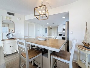 Ein Essbereich mit einem Holztisch und weißen Stühlen, einer Deckenleuchte und einer Küche im Hintergrund.