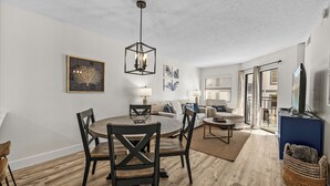 A modern living room with a dining area, featuring a round table, dark wooden chairs, a sofa, and a TV stand.