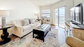 Ein modernes Wohnzimmer mit einem weißen Sofa, einem schwarzen Hocker, einem Fernseher und einem großen Fenster mit Blick auf die Stadt.