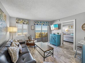 Un salon avec un canapé, une table basse et un téléviseur fixé au mur. Il y a un balcon avec vue sur la plage.