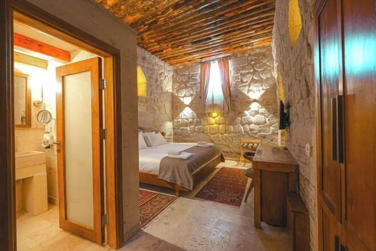 Un dormitorio con pared de piedra, techo de madera, una cama, una mesita de noche y un pequeño armario de madera.