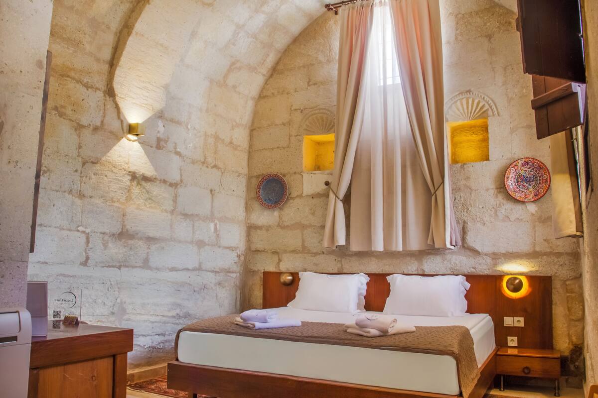 Una habitación con paredes de piedra, una cama, lámparas de noche y un ventanal con cortina.