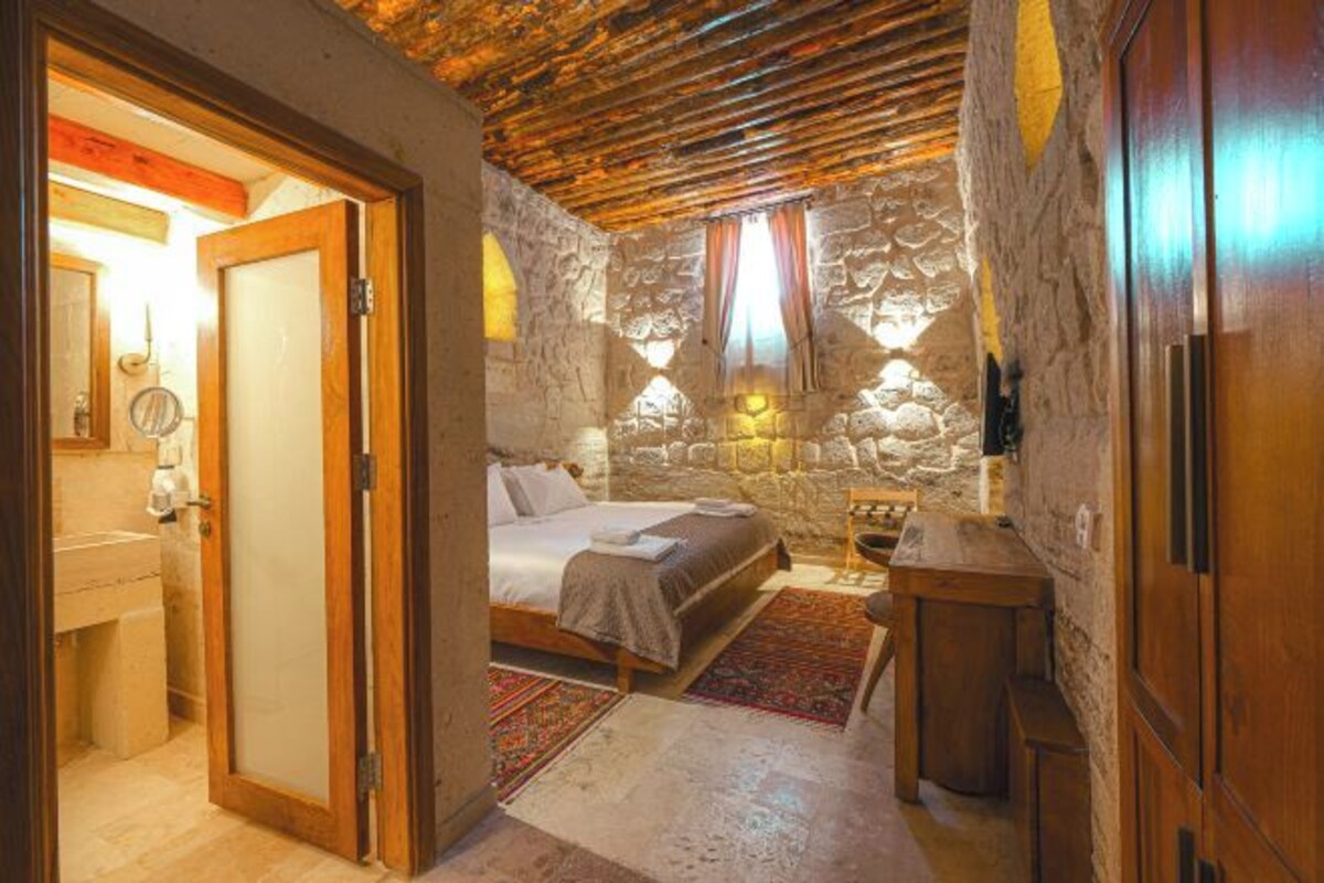 Une chambre avec un mur en pierre, un plafond en bois, un lit, une table de chevet et une petite armoire en bois.