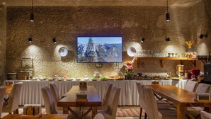 Un espace repas avec une longue table en bois, des chaises blanches, un buffet garni de divers plats et un téléviseur fixé au mur.