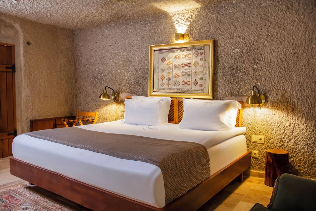 Un dormitorio con una cama grande, un cabecero de madera, una obra de arte enmarcada sobre la cama y dos lámparas fijadas a la pared.