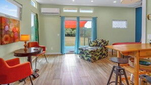 A room with a dining table, two chairs, a sofa, and a stool. There is a window with curtains, an air conditioning unit, and a door leading outside.