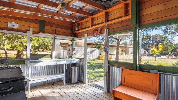 A covered outdoor area with a wooden deck, a grill, a bench, and a view of a garden.