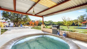 A covered outdoor swimming pool with a hot tub, a barbecue grill, and a view of a backyard with trees and a lawn. - The Main House at Starlight Meadow (Wimberley)