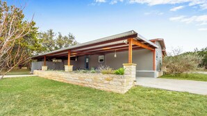 A modern house with a covered patio, stone retaining wall, and a clear blue sky. - The Main House at Starlight Meadow (Wimberley)