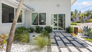 Een modern huis met een stenen pad dat leidt naar een glazen deuropening, omgeven door diverse planten en een palmboom.