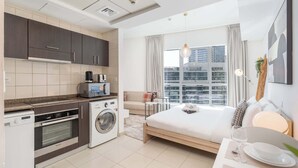 A modern hotel room with a kitchenette, a bed, a dining area, and a large window with a city view.