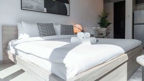 A modern bedroom with a large bed, a wall-mounted abstract painting, a bedside lamp, and a small table with a plant.