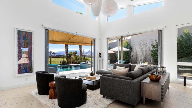 A spacious living room with a large sofa, black chairs, a coffee table, and a view of a swimming pool and mountains.