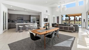A modern kitchen with a central island, a dining area with a wooden table and chairs, and a living area with a sofa and a view of the outdoors.