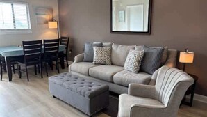 Un salon avec un canapé gris, un pouf assorti et un coin repas comprenant une table et des chaises.