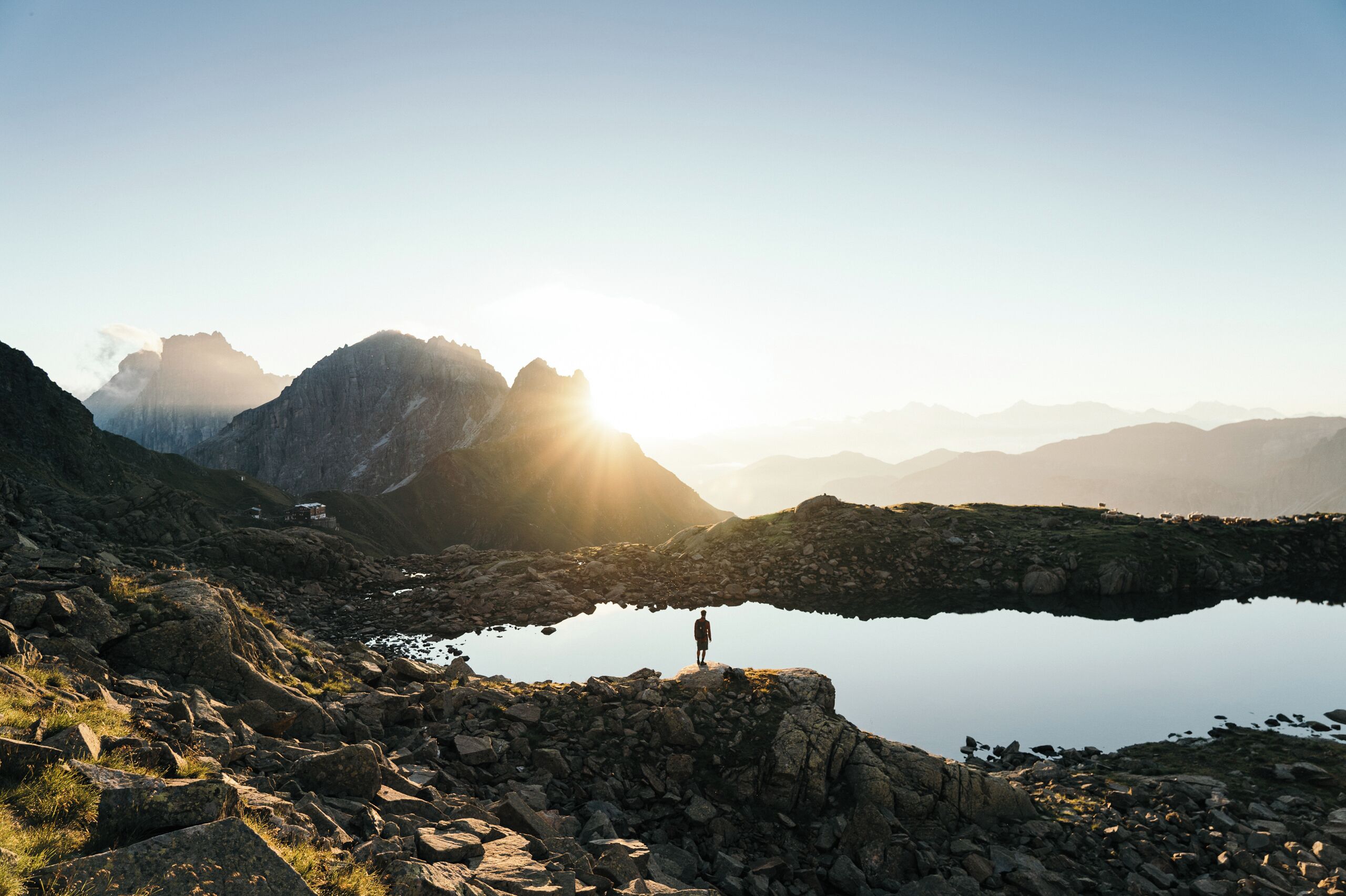 Una persona está de pie sobre un saliente rocoso, contemplando un lago tranquilo con montañas al fondo.