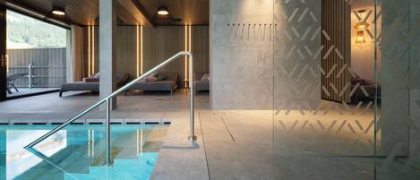 A modern indoor pool area with a glass barrier, a metal staircase, and a seating area with cushions.