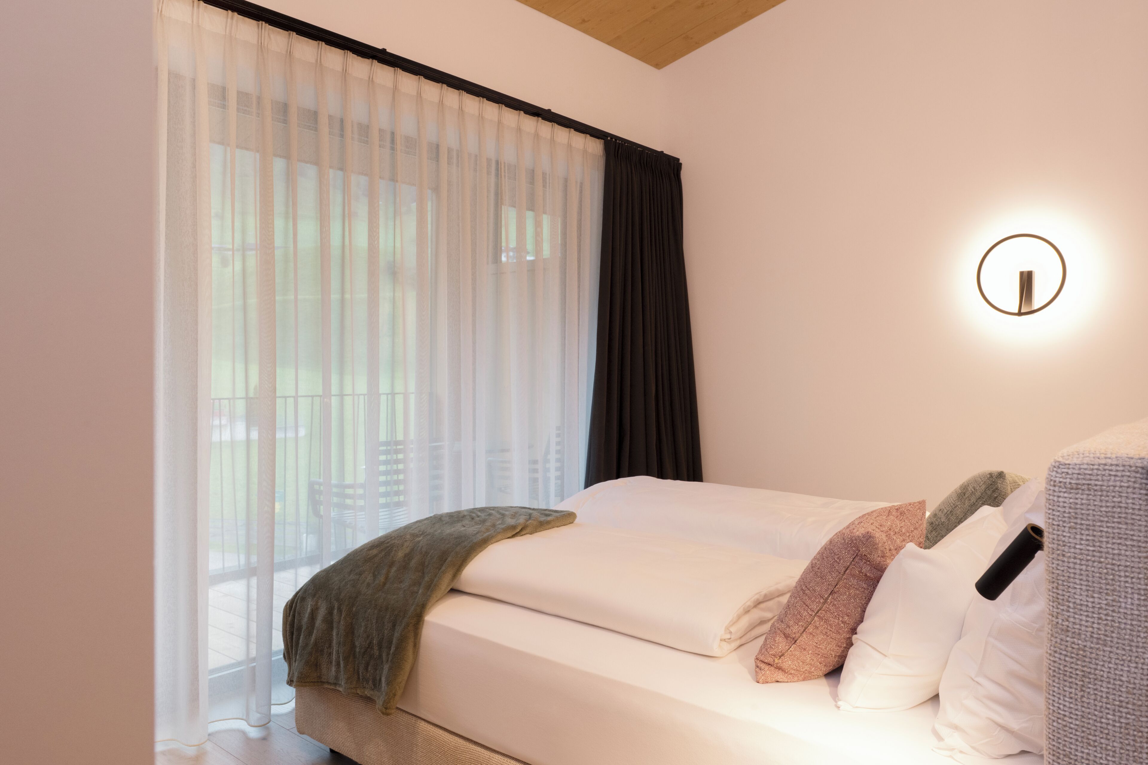 A bedroom with a bed, a window with sheer curtains, a dark curtain, and a wall-mounted light fixture.