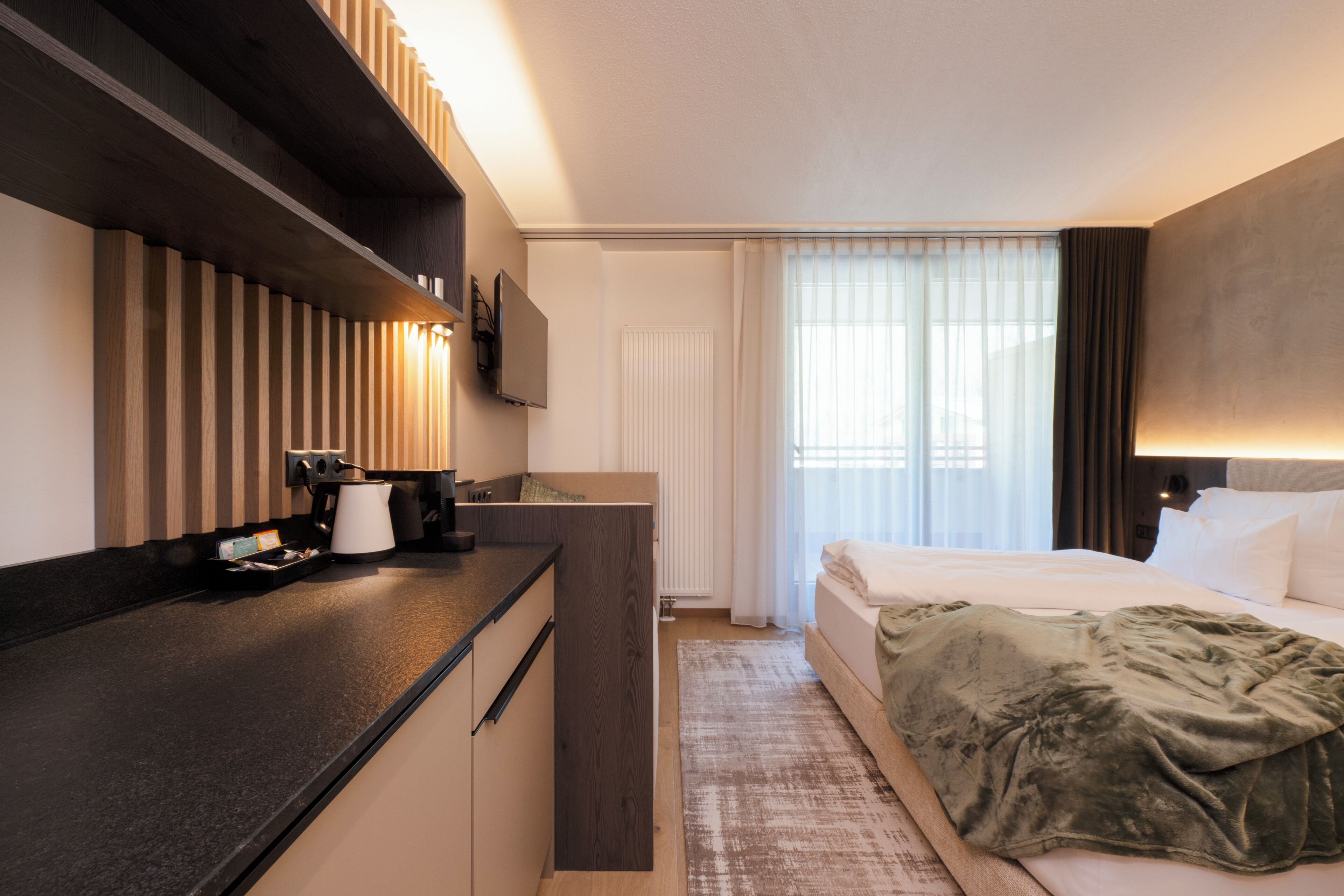 A modern hotel room with a kitchenette, a bed with a green throw, a large window with curtains, and a flat-screen TV mounted on the wall.