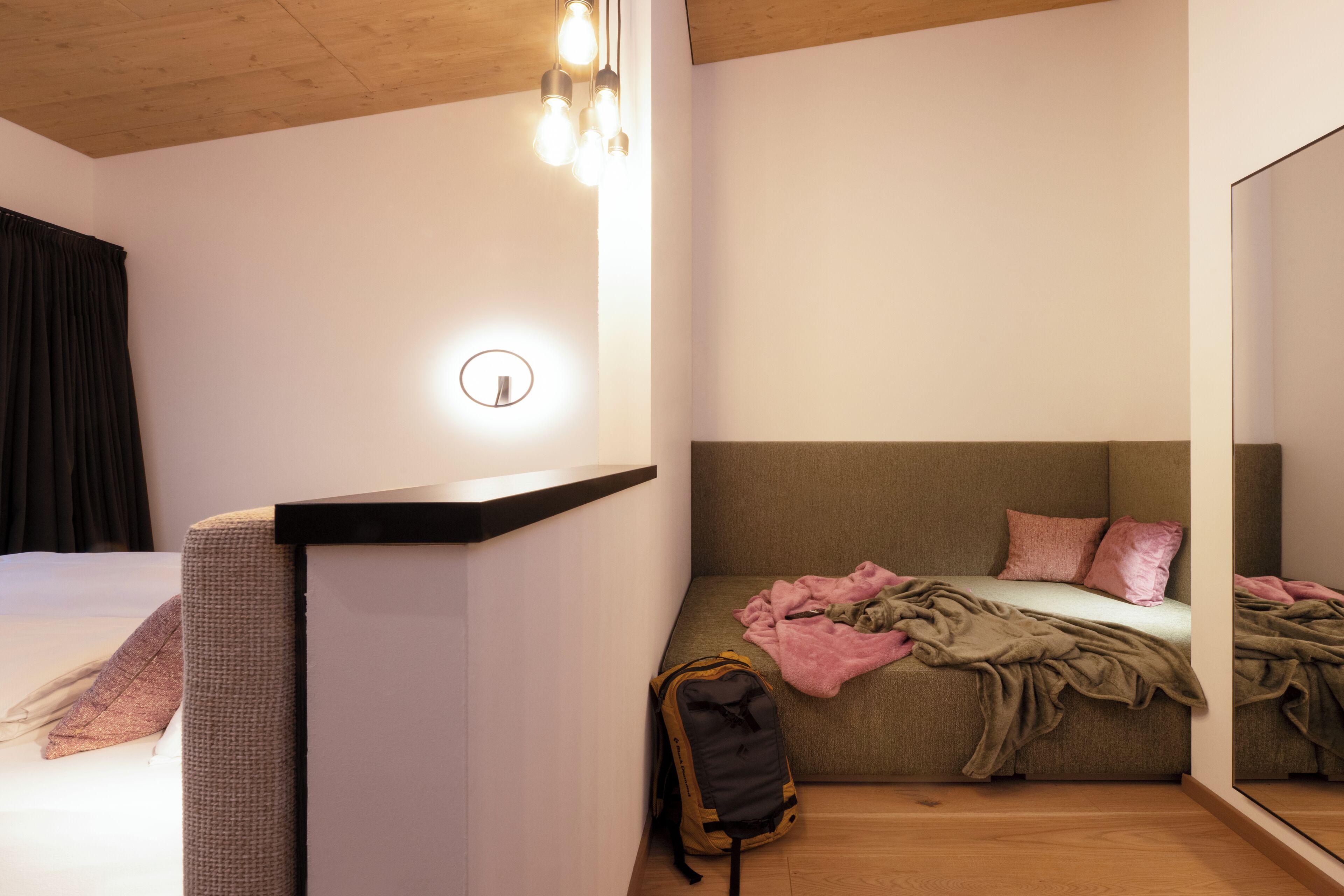 A bedroom with a bed, a bedside table, a mirror, a wall-mounted light, and a backpack on the floor.