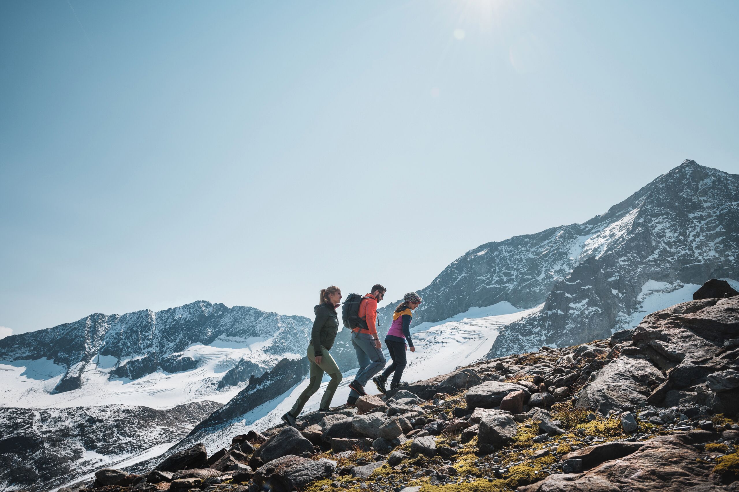 Three hikers on a rocky mountain trail with snow-covered peaks in the background.