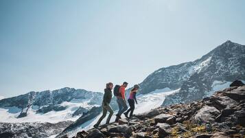 Three hikers on a rocky mountain trail with snow-covered peaks in the background.
