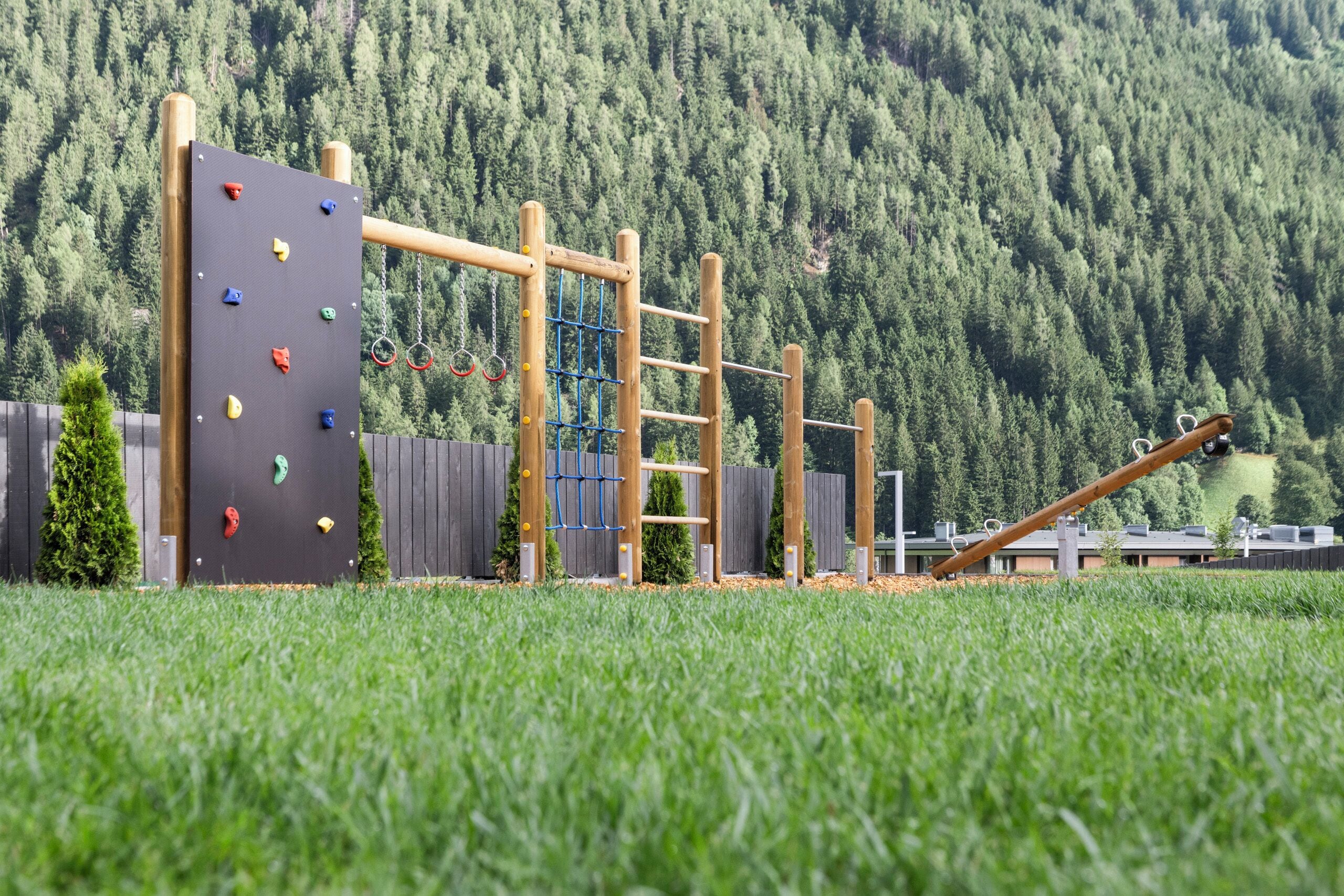 Une aire de jeux dotée d’installations d’escalade et de jeux suspendus, entourée d’une pelouse et bordée, au loin, par une colline boisée.